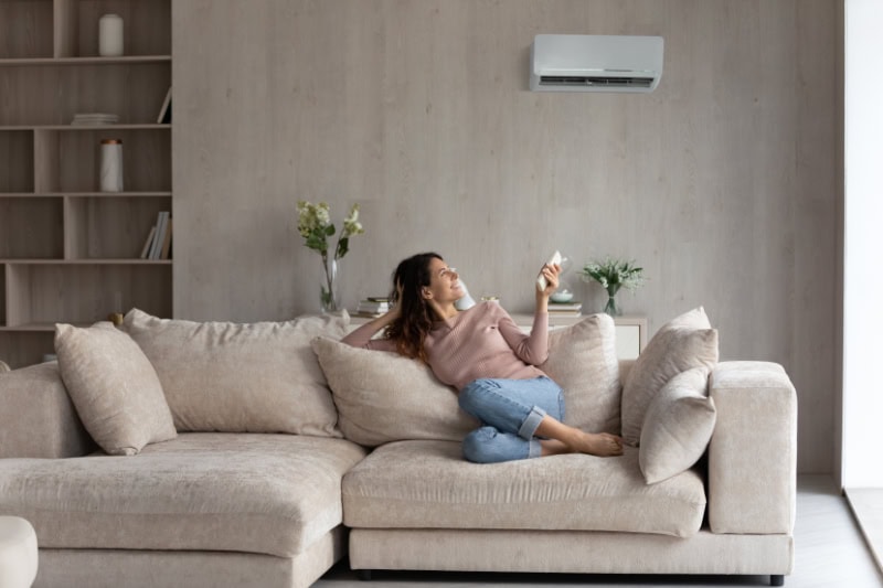 Planning to Remodel? Go Ductless! Relaxed young Hispanic female homeowner sitting on huge comfortable couch, turning on air conditioner with remote controller, switching on cooler system, setting comfortable temperature in living room.