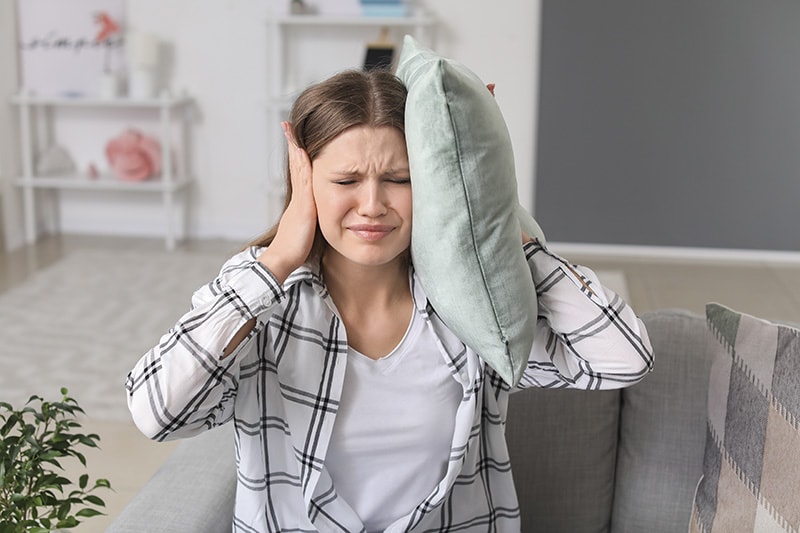 A young woman covering her ears with a pillow from loud noise, Why Is My Heat Pump Making Loud Noises? | HVAC Maintenance | Dana's