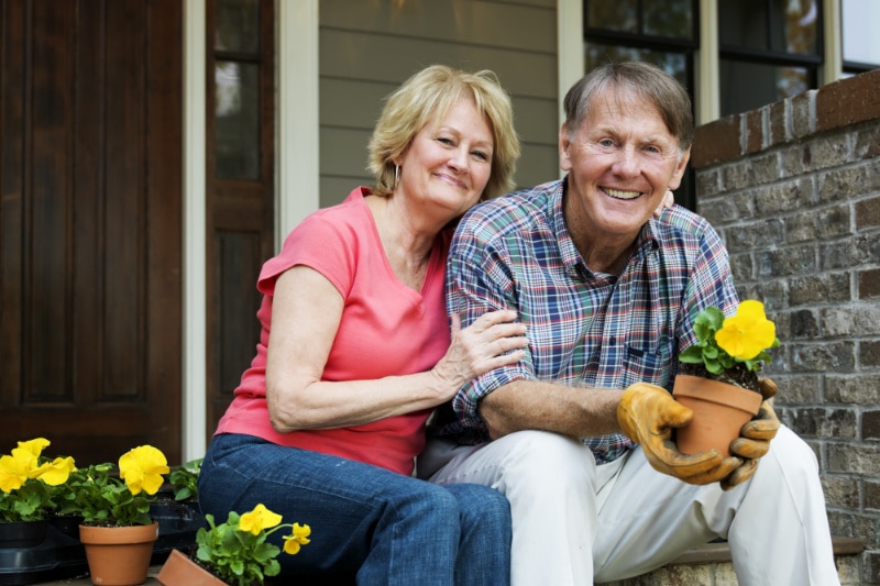 Senior Man And Woman Sit Outside Their House And Smile At The Camera Holding Pot Plant.