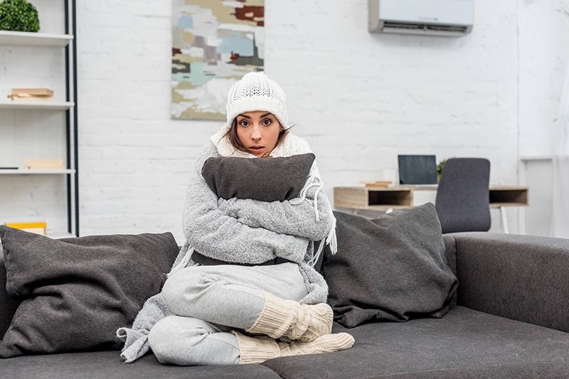 freezed young woman in warm clothes sitting on couch and hugging cushion at home.