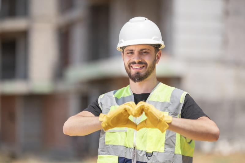 How to Join the HVAC Industry. Image is a photograph of an HVAC professionals wearing a white helmet and safety vest and making a heart with his hands.